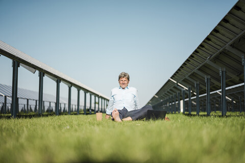 Älterer Mann sitzt barfuß auf einer Wiese, Laptop und Coffee-to-go-Becher, Solaranlage, lizenzfreies Stockfoto