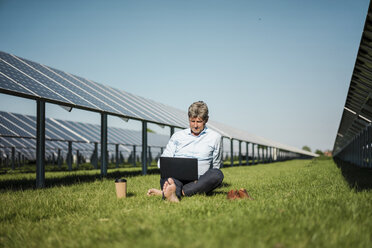 Älterer Mann sitzt barfuß auf einer Wiese, Laptop und Coffee-to-go-Becher, Solaranlage - MOEF01142