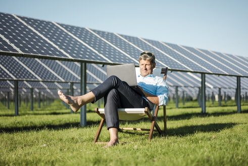 Mature man sitting in beach lounger, using laptop, solar plant - MOEF01155