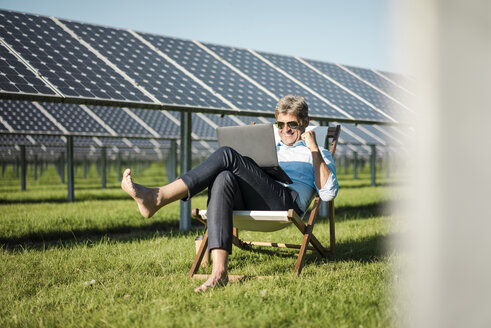 Mature man sitting in beach lounger, using laptop in solar plant - MOEF01156