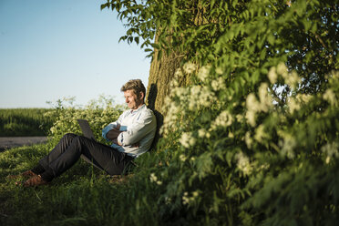 Geschäftsmann lehnt an einem Baum und benutzt einen Laptop - MOEF01203