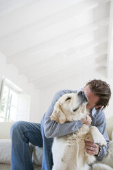 Junger Mann auf dem Wohnzimmersofa, der einen Golden Retriever Hund umarmt - CUF13695