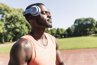 Young athlete exercising on sports field, eyes close, wearing headphones - UUF13902