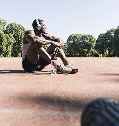 Junger Sportler sitzt auf einem Sportplatz und hört Musik - UUF13912