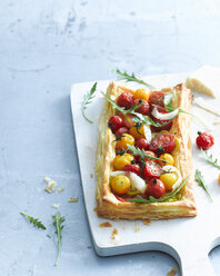 Draufsicht auf eine Blätterteigtorte mit Tomaten und Parmesan, garniert mit Rucola auf einem Schneidebrett - CUF13881