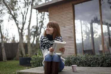 Woman sitting on picnic table holding book looking away smiling - CUF14121