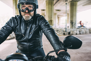 Älterer männlicher Motorradfahrer mit schwarzem Helm und Schutzbrille auf dem Motorrad sitzend unter einer Überführung - CUF14462