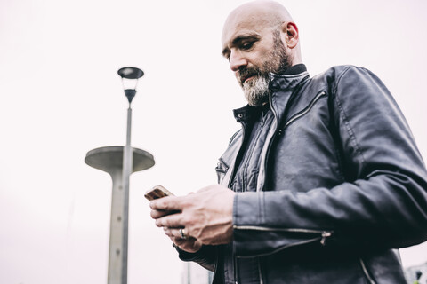 Älterer männlicher Motorradfahrer, der vor grauem Himmel einen Text auf seinem Smartphone liest, lizenzfreies Stockfoto