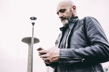 Älterer männlicher Motorradfahrer, der vor grauem Himmel einen Text auf seinem Smartphone liest - CUF14477