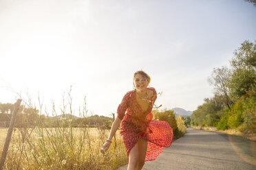 Glückliche junge Frau in rotem Kleid beim Laufen auf einer Landstraße, Mallorca, Spanien - CUF14488