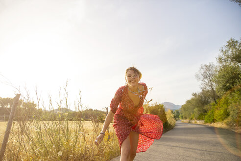 Glückliche junge Frau in rotem Kleid beim Laufen auf einer Landstraße, Mallorca, Spanien - CUF14488