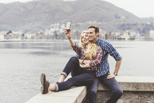 Junges Paar sitzt an der Hafenmauer und macht ein Smartphone-Selfie, Comer See, Italien - CUF14618