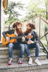 Junge männliche Hipster-Zwillinge mit roten Bärten sitzen auf einer Treppe und spielen Gitarre - CUF14649