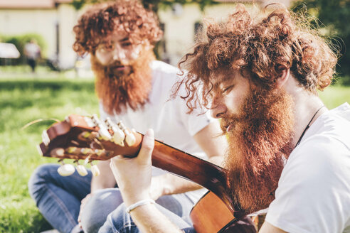 Young male hipster twins with red beards sitting in park playing guitar - CUF14651