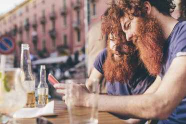 Junge männliche Hipster-Zwillinge mit roten Haaren und Bärten lesen Smartphone-Texte in einer Bar am Straßenrand - CUF14655