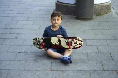 Porträt eines staunenden kleinen Jungen, der mit einem Skateboard auf dem Bürgersteig sitzt, lizenzfreies Stockfoto