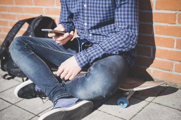 Blick von unten auf einen jungen männlichen Skateboarder, der auf dem Gehweg sitzt und einen Text auf seinem Smartphone liest - CUF14844
