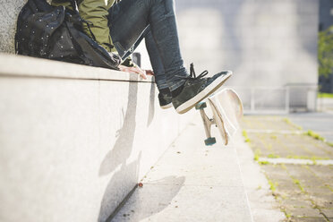 Blick von der Taille abwärts auf einen jungen männlichen Skateboarder, der auf einer Wand sitzt und mit den Füßen ein Skateboard umdreht - CUF14863