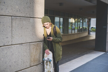 Young male urban skateboarder leaning against wall reading smartphone texts - CUF14869