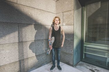 Portrait of young male urban skateboarder standing in corner holding skateboard - CUF14870