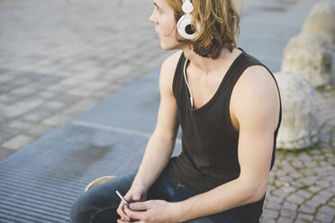 Junger Mann sitzt auf dem Bürgersteig und hört über Kopfhörer Musik - CUF14874