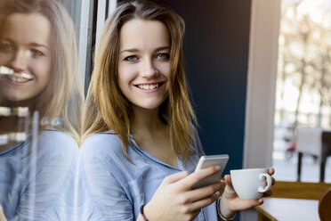 Young woman holding coffee cup and smartphone looking at camera smiling - CUF15092