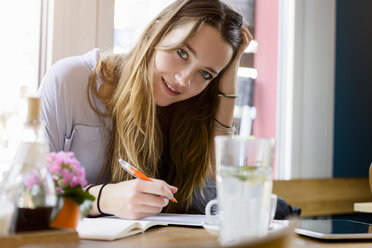 Junge Frau im Café beim Schreiben, die Hand auf den Kopf gestützt, lächelnd in die Kamera blickend - CUF15099