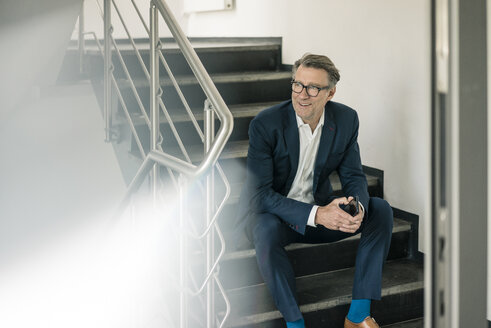 Smiling mature businessman with cell phone sitting on stairs - JOSF02186