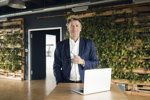 Porträt eines selbstbewussten reifen Geschäftsmannes mit Laptop in einem grünen Büro, lizenzfreies Stockfoto
