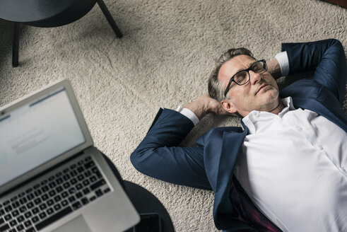 Älterer Geschäftsmann auf Teppich liegend neben einem Laptop - JOSF02199