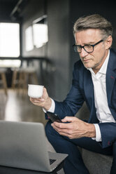 Mature businessman with cup of coffee and laptop using cell phone - JOSF02201