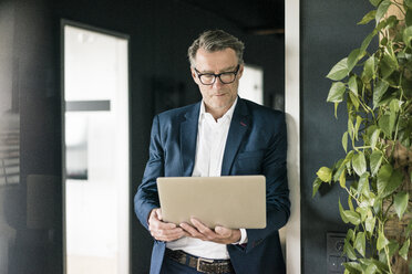 Mature businessman standing in office using laptop - JOSF02206