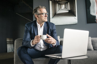 Älterer Geschäftsmann sitzt auf der Couch mit einer Tasse Kaffee und einem Laptop - JOSF02263