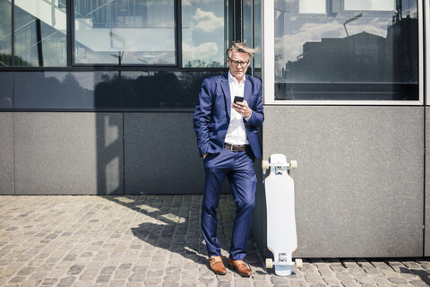 Älterer Geschäftsmann benutzt Handy im Freien neben Longboard, lizenzfreies Stockfoto