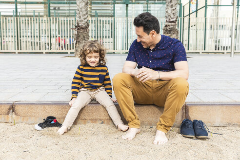 Spain, Barcelona, father and son sitting next to the beach barefoot - WPEF00353