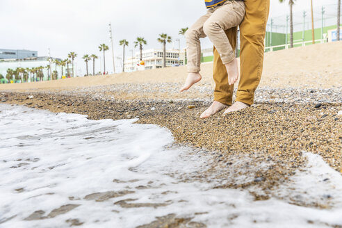 Spain, Barcelona, father holding his son on the beach next to waves - WPEF00367