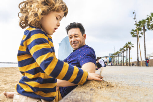 Spain, Barcelona, young boy playing with sand, his father sitting next to him and smiling - WPEF00369