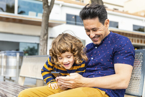 Spain, Barcelona, happy father and son with a smartphone sitting on bench - WPEF00377