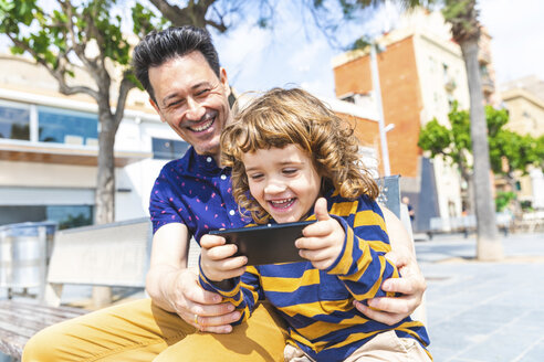 Spanien, Barcelona, glücklicher Vater und Sohn mit einem Smartphone auf einer Bank sitzend - WPEF00379