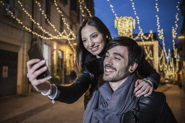 Junger Mann, der eine junge Frau huckepack nimmt und dabei sein Smartphone benutzt, um ein Selfie zu machen, lächelnd - CUF15179