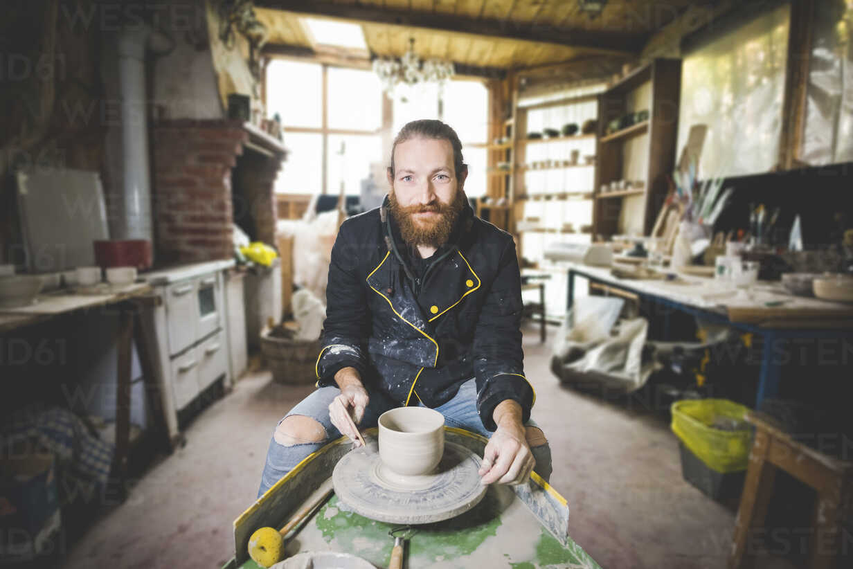bearded-mid-adult-man-in-workshop-sitting-at-pottery-wheel-making-clay-pot-looking-at-camera-smiling-CUF15228.jpg