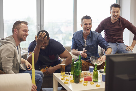 Eine Gruppe von Männern sieht sich ein Sportereignis im Fernsehen an, während sie lächelnd Snacks und Biere genießen, lizenzfreies Stockfoto