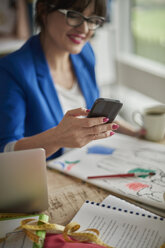 Frau im Designstudio sitzt am Schreibtisch und schaut lächelnd auf ihr Smartphone - CUF15453