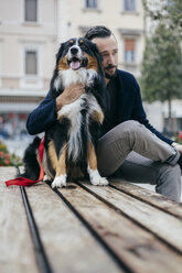 Mid adult man sitting with pet dog on city square bench - CUF15681