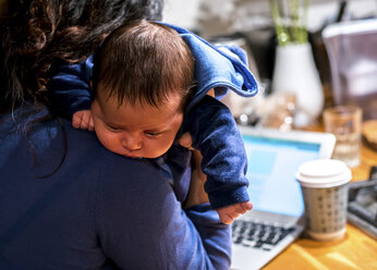 Mother cradling newborn son and using laptop on kitchen bar - CUF15710