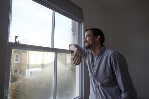 Mittlerer erwachsener Mann, der durch ein Schlafzimmerfenster hinausschaut, lizenzfreies Stockfoto
