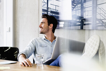 Business man in office feet up on desk, using laptop looking away smiling - CUF15758