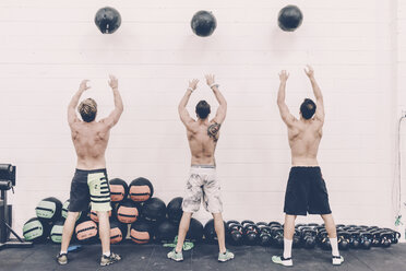 Rückansicht von drei männlichen Crosstrainern, die in einem Fitnessstudio Übungsbälle werfen - CUF15876