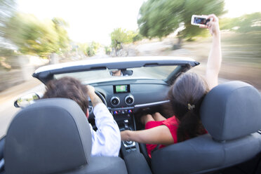 Rückansicht eines jungen Paares, das ein Selfie macht, während es auf einer Landstraße im Cabrio fährt, Mallorca, Spanien - CUF16101