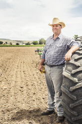 Portrait of male farmer leaning against tractor tyre in ploughed field - CUF16161